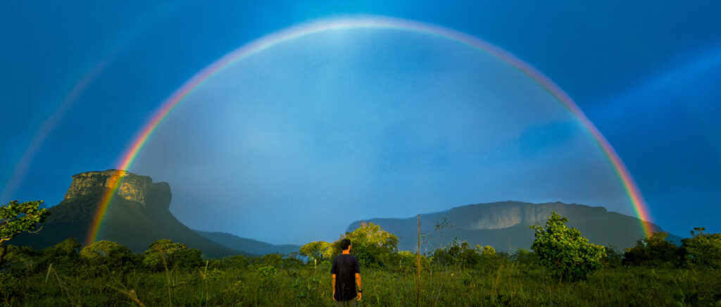 Person contemplates a full rainbow with Camel Hill in the background, in Chapada Diamantina.