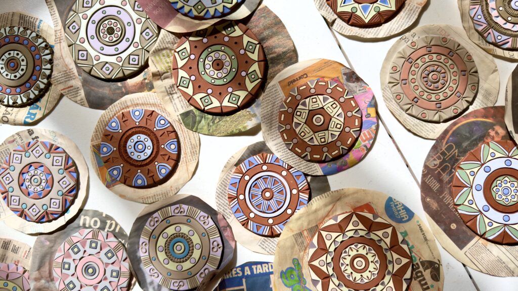 Mandalas circulares em cerâmica feitas por Silvia Lopes, dispostas sobre recortes de jornal. As peças têm desenhos geométricos em relevo, tons terrosos e detalhes coloridos em azul, rosa, verde e branco.