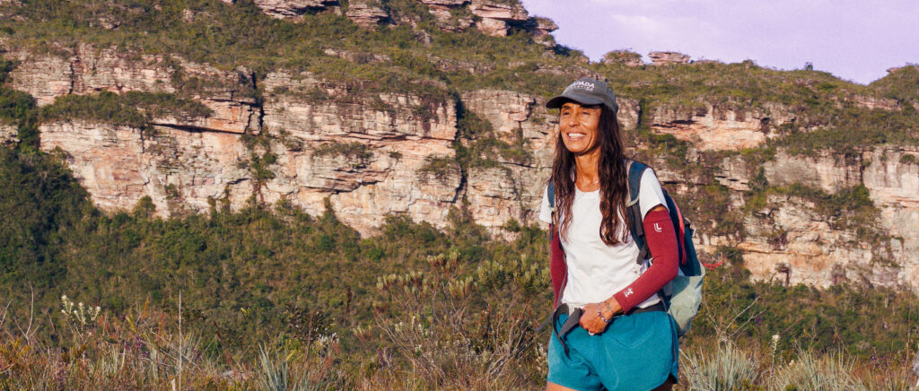 Guia Cris Pascoli caminhando por uma trilha do Vale do Pati, com formação rochosa da Serra do Sincorá ao fundo, na Chapada Diamantina.